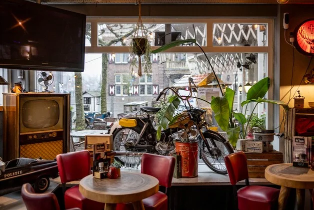 Interieur van De Zaak met rode stoelen, een oude brommer en retro-tv bij het raam